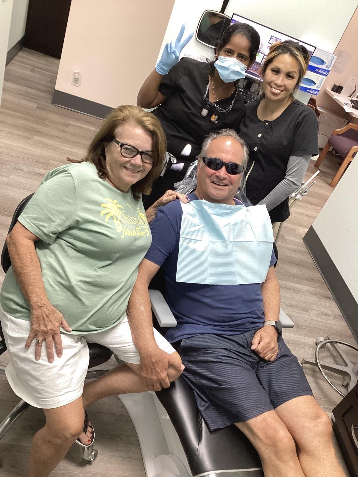 A man is sitting in a dental chair with two women standing around him.