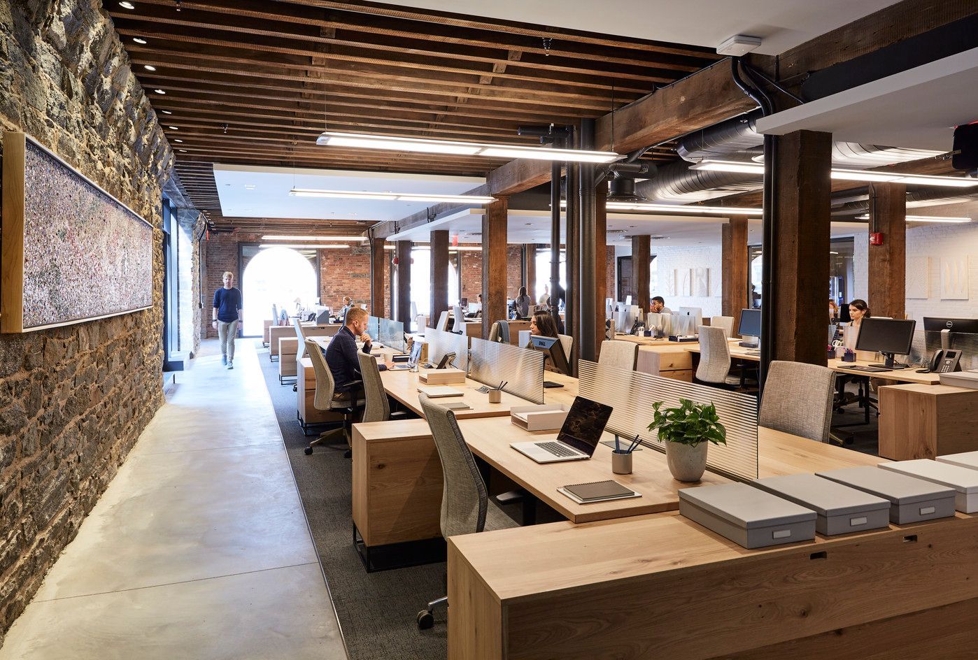 Open-plan office with wood desks, stone walls, exposed beams, and people working on computers.