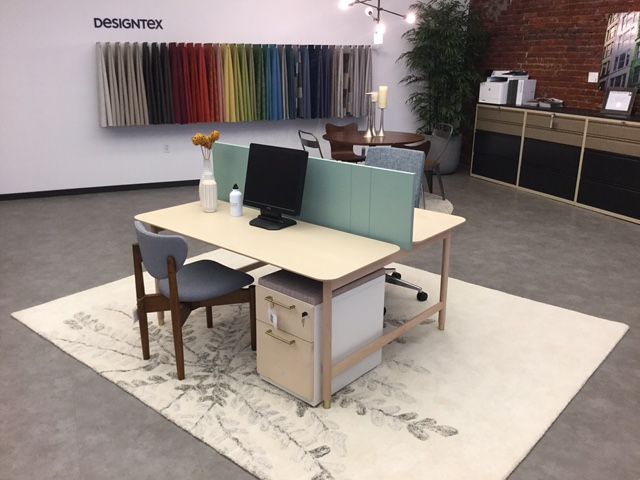 Office space with desks, fabric samples, and a rug. Light wood desk with computer, light green dividers, and a grey chair.