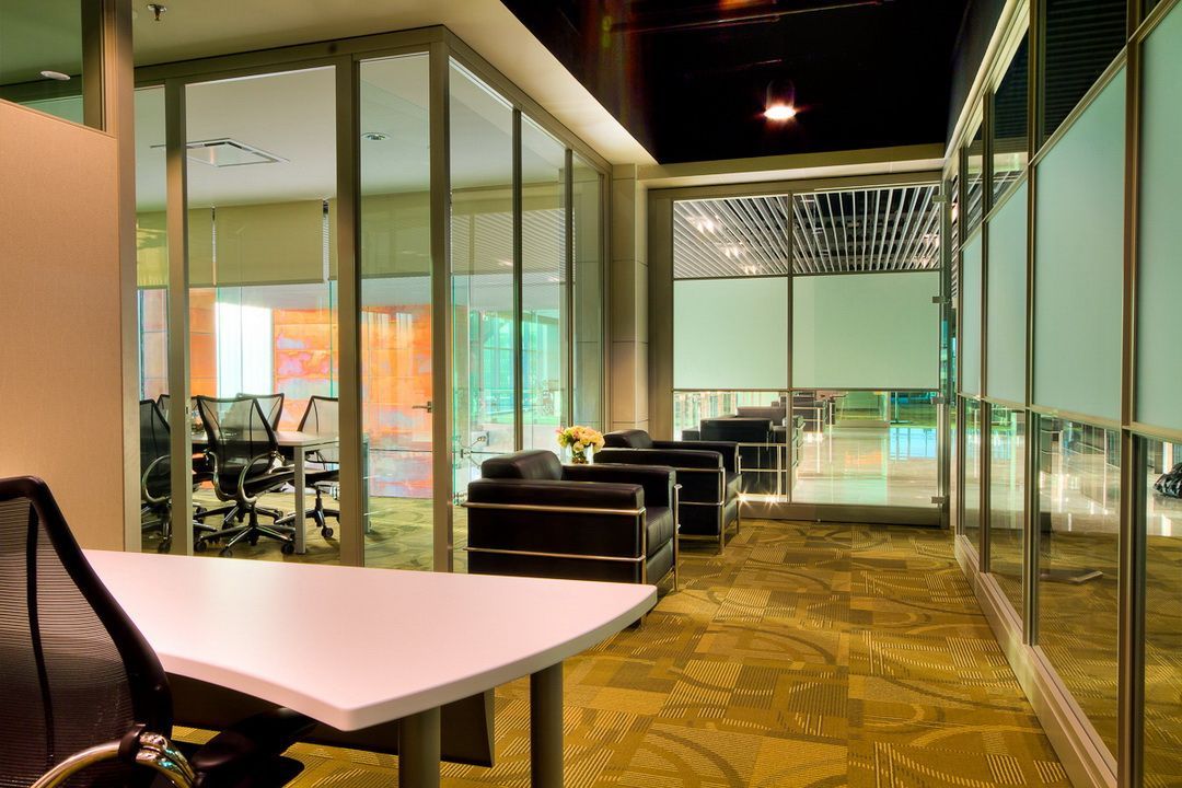 Modern office interior with glass-walled cubicles, waiting area with black chairs, and patterned carpet.