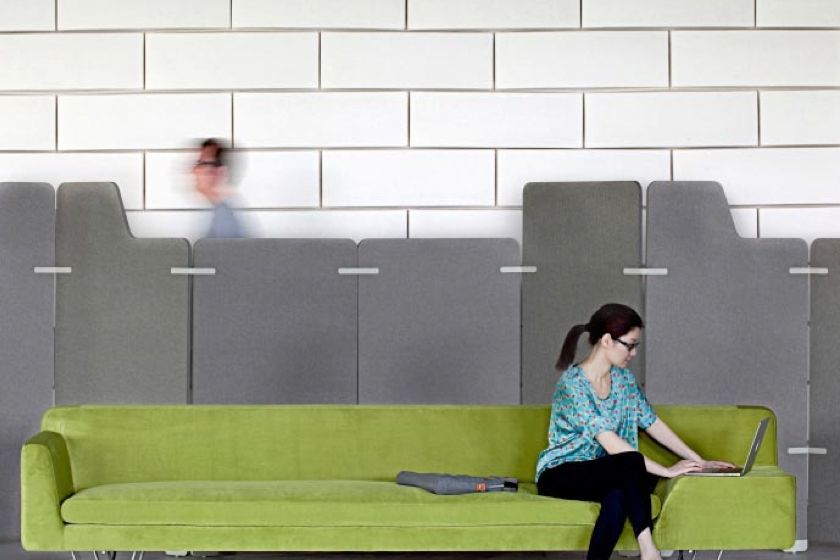 Woman on a green sofa working on a laptop, in front of gray dividers and a white brick wall.