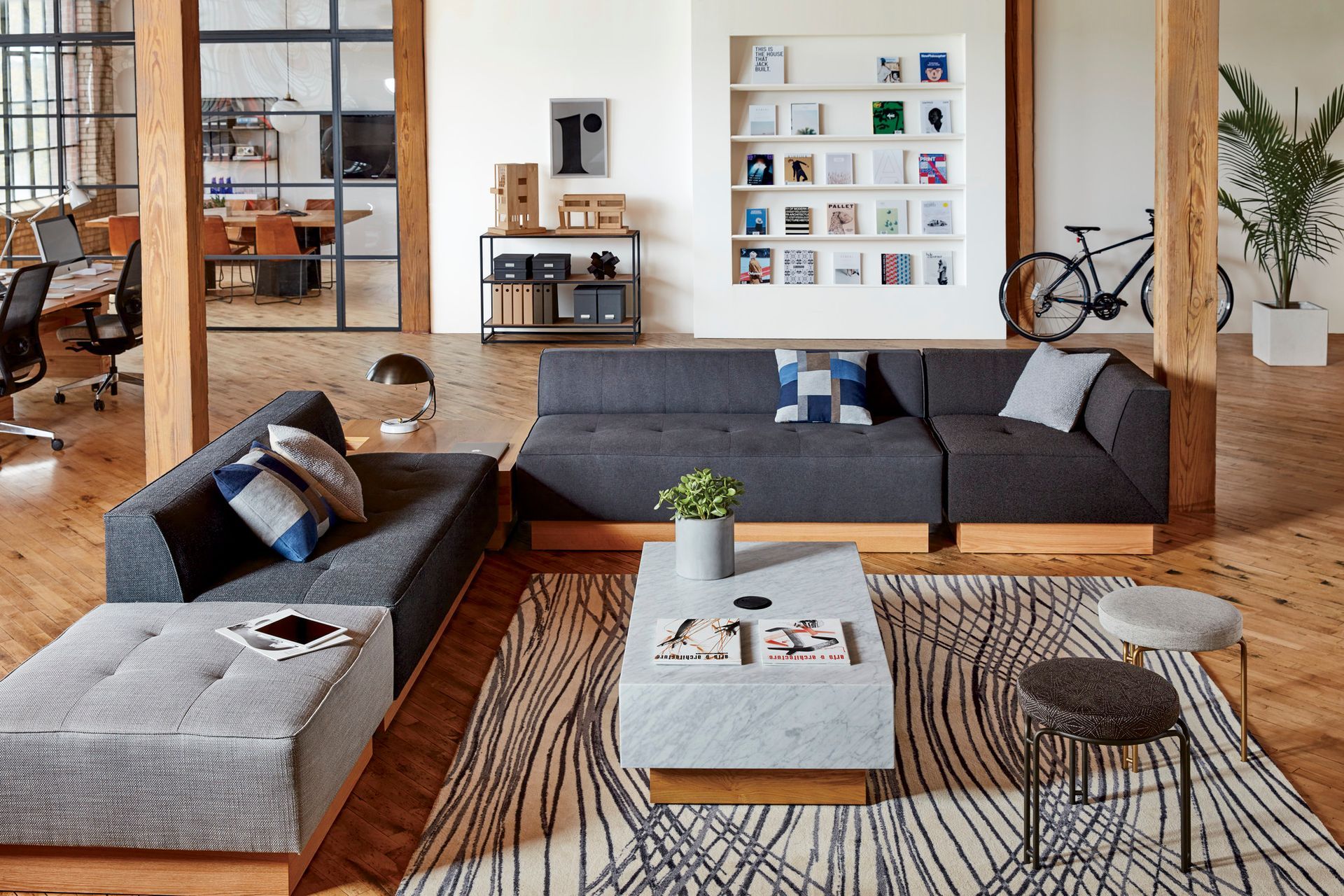 Modern office lounge area with dark gray sectional sofa, patterned rug, marble coffee table, and bicycle.