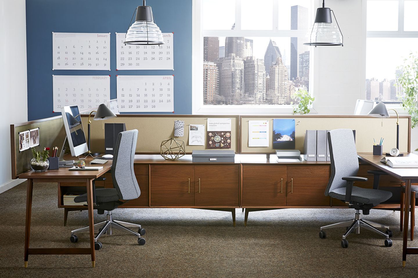 Office space with wooden desks, chairs, and cabinets. Calendars, lamps, and city view through the window.