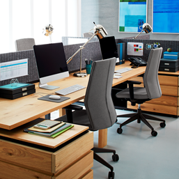 Office space with light wood desks, gray chairs, monitors, lamps, and artwork.