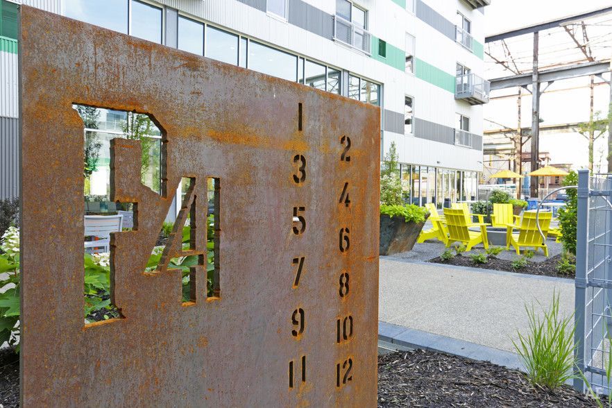 Rusty metal sign with the numbers 34 and a vertical row of numbers 1-12 in front of a modern building.
