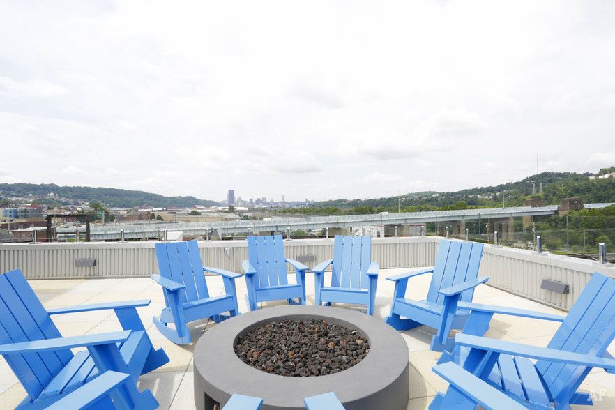 Blue Adirondack chairs circle a fire pit on a rooftop, overlooking a city and a bridge under a cloudy sky.