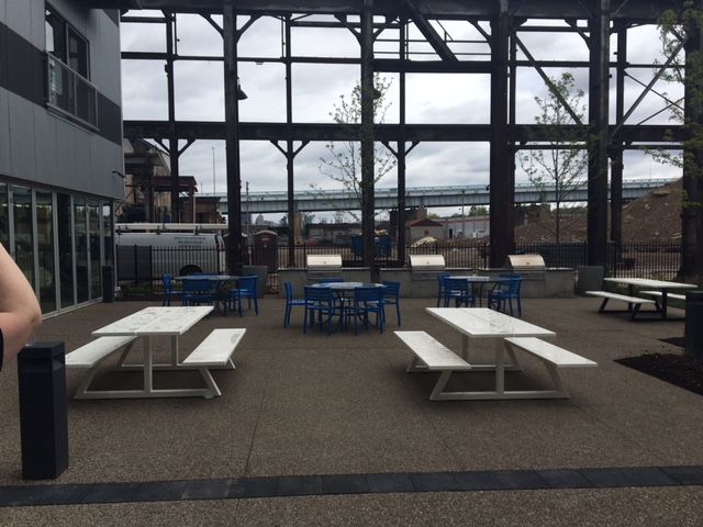 Outdoor patio with picnic tables, blue chairs, and open-air structure. Bridge visible in the background.