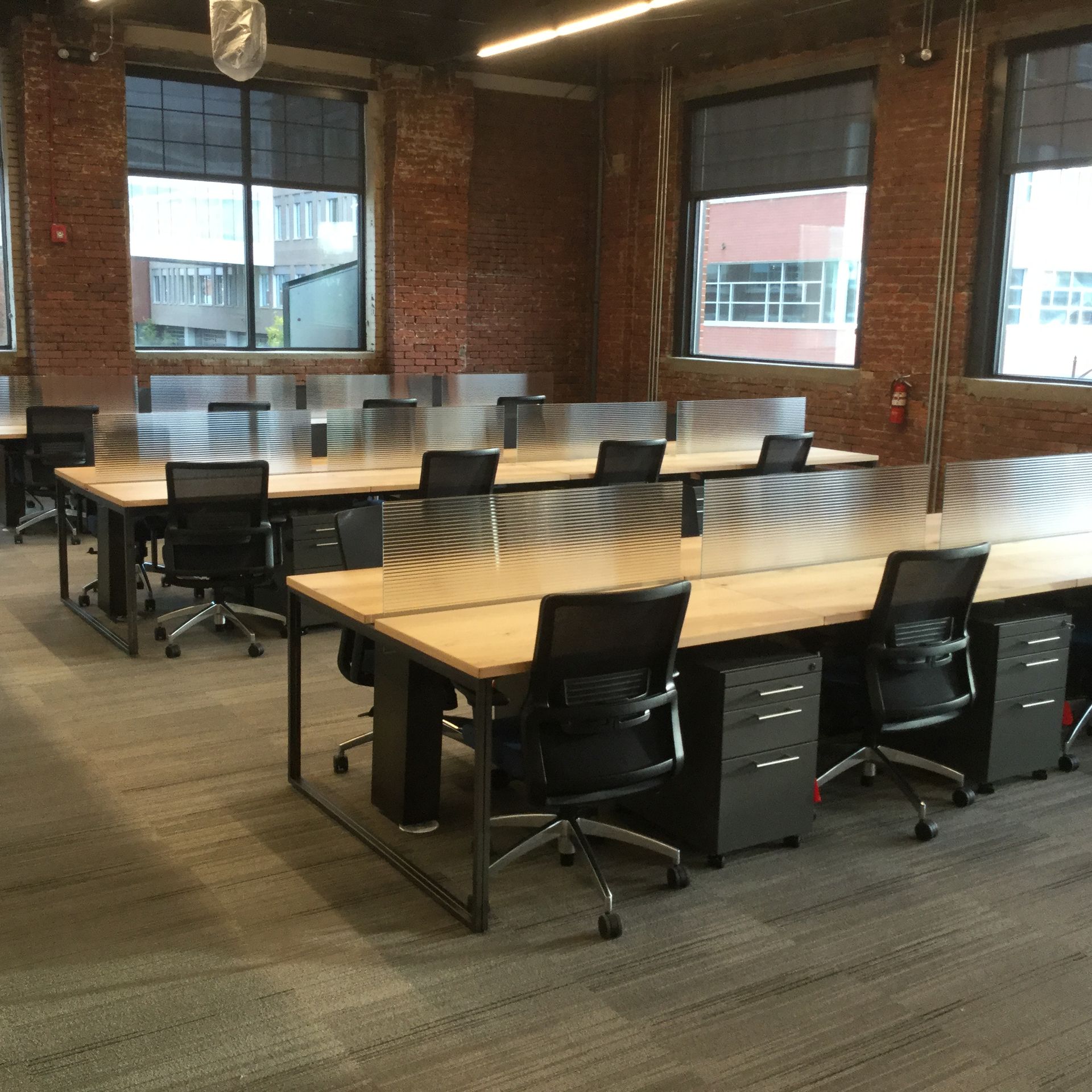 Open office with desks, chairs, metal dividers, brick walls, and windows.