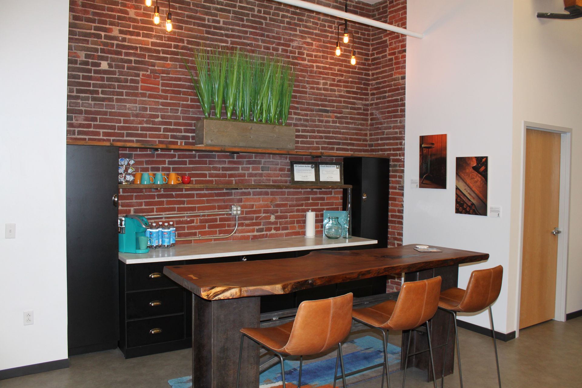 Loft-style kitchenette with brick wall, wooden bar, leather stools, and a black cabinet.