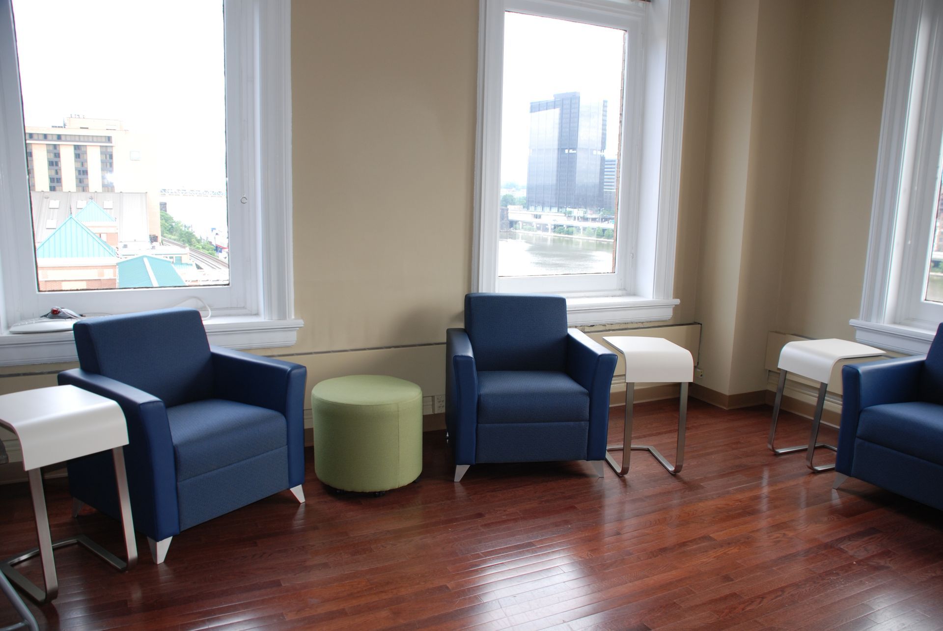 Three blue armchairs, small white side tables, and a green ottoman in a room with large windows overlooking a city.