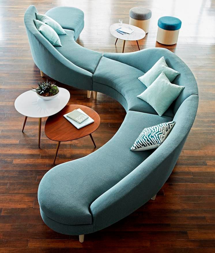 Curved teal sofa with pillows, surrounded by small round tables, on a wood floor.