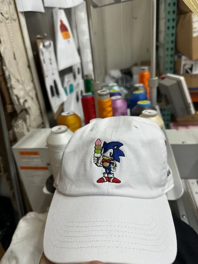 White baseball cap with embroidered Sonic the Hedgehog holding an ice cream cone, set in a workshop.