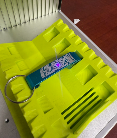 Teal keychain with white text and a silver ring on a yellow textured surface, lit by a purple glow.