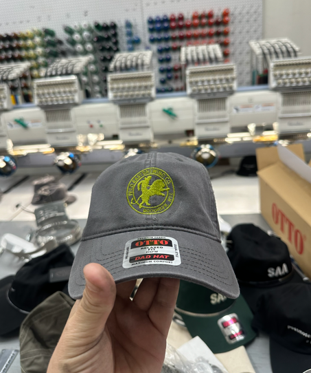 Person holding a gray baseball cap with embroidered logo, in front of embroidery machines.
