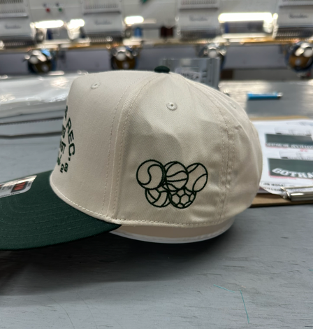Cream and green baseball cap with embroidered sports balls, in a factory setting.