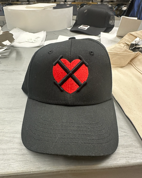 Black baseball cap with a red heart-shaped logo crossed by black lines.