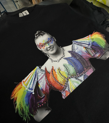 Black t-shirt with a colorful printed image of a person smiling, wearing a jeweled bodice and rainbow sleeves.