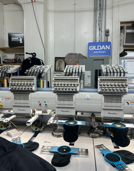 Three embroidery machines in a workshop with a Gildan sign. Black thread spools and blue accessories.