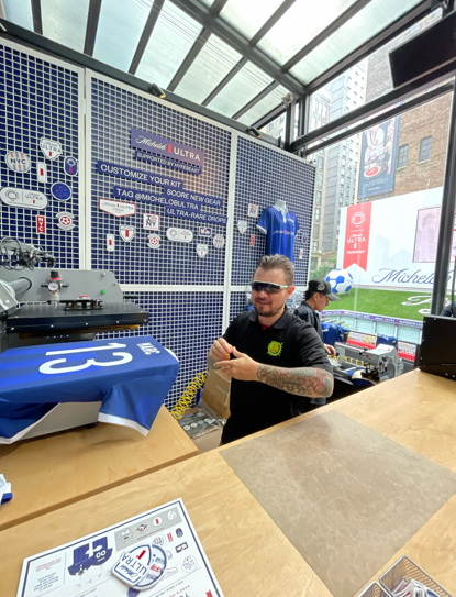 Man at a booth printing a jersey; blue, white, and wood tones; Chelsea FC branding.