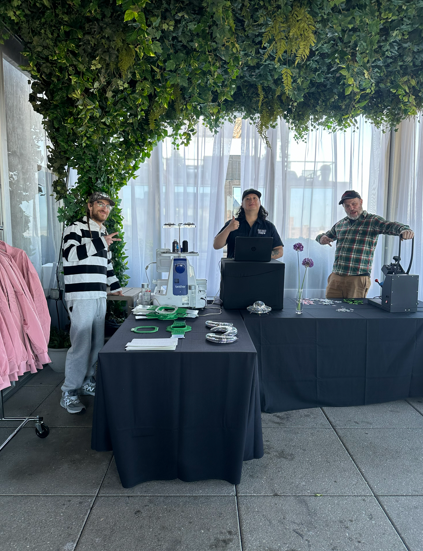 Three people stand behind tables at an outdoor event. Greenery hangs overhead.