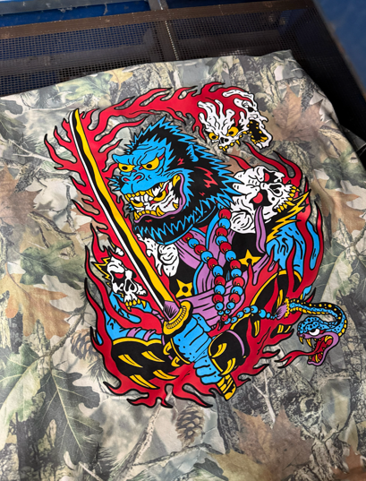 Vibrant illustration of a blue-faced demon warrior with a sword, surrounded by red flames and skulls, on camo fabric.