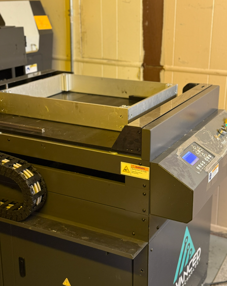 A black industrial printer with a metal tray. The printer is in a room with a light-colored wall.