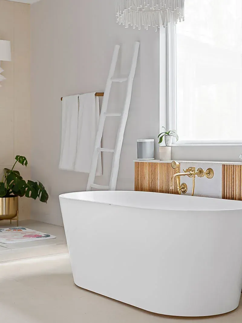 White bathtub in a bright bathroom with a ladder, gold fixtures, and a window.