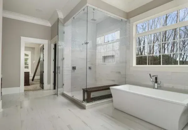 Modern bathroom with glass shower, white tub, and large window overlooking trees.