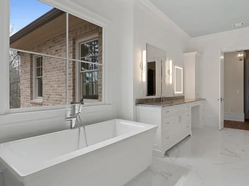 Bright white bathroom with a tub, vanity, and large window overlooking a brick exterior.
