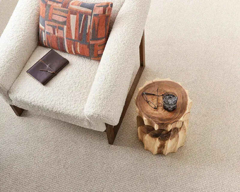 Cozy living area with a white armchair, patterned pillow, and wooden side table on beige carpet.
