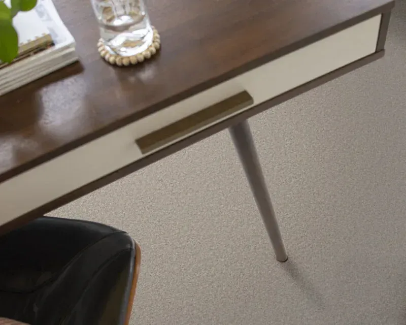 Wooden desk with white drawer, brass handle, and light gray leg on a speckled carpet.