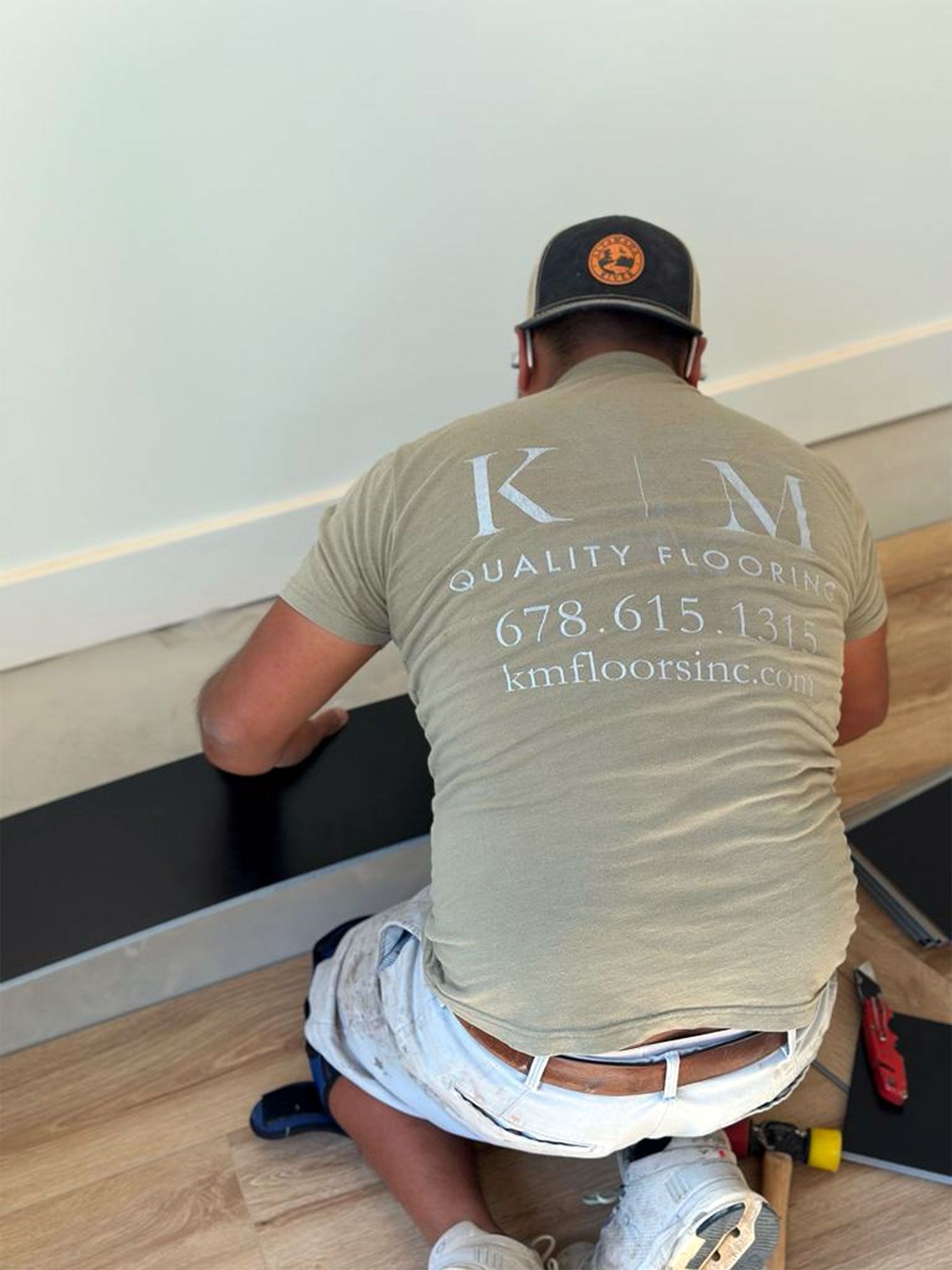 Person installing black flooring near a white wall. Beige shirt with 