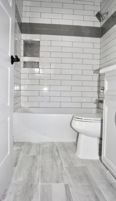 Bathroom with white and gray tiled walls, tub, toilet, and gray wood-look floor.