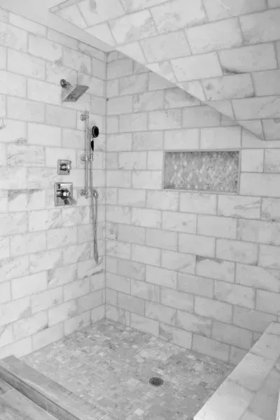 Shower with white marble tile, showerhead, handheld sprayer, and recessed shelf.