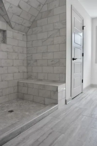 A tiled shower with a built-in seat and arched ceiling. Light gray tiles, white door in the background.