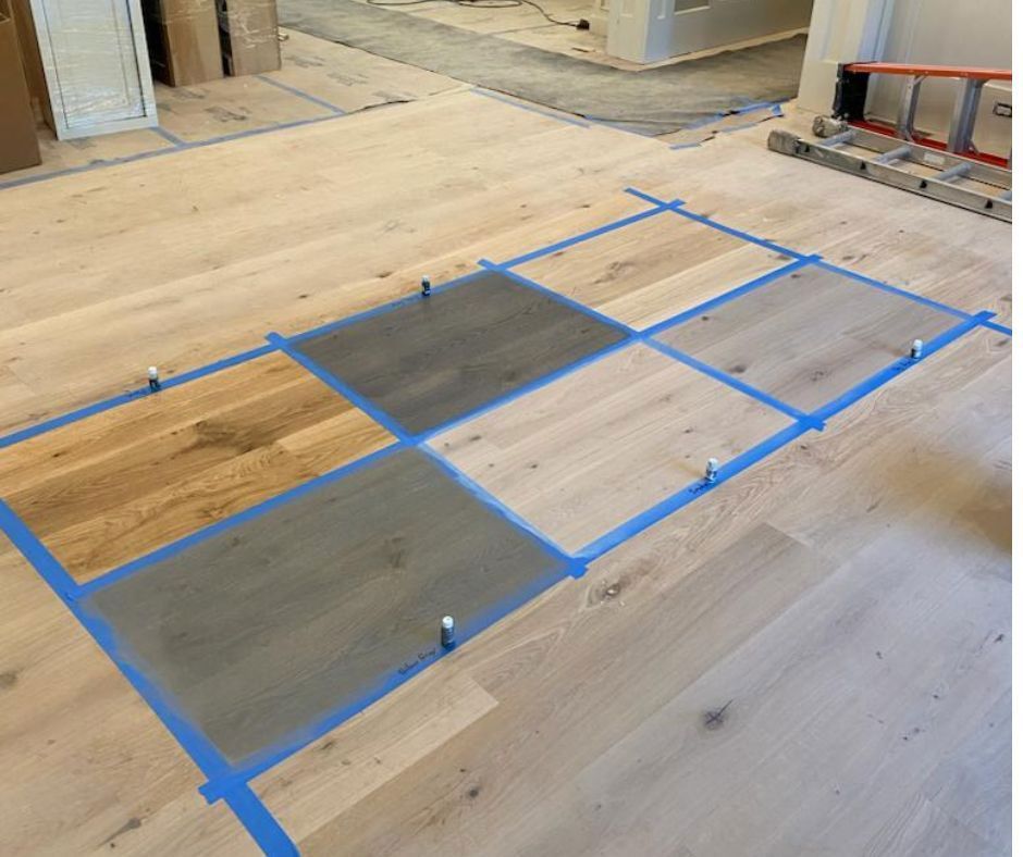 Floor samples on hardwood flooring, with blue tape outlining. Some samples are stained gray, others natural wood tone.