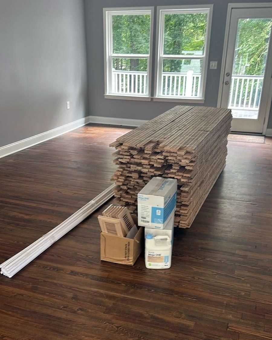Room with wood flooring and stacked planks, boxes, and trim in front of windows.