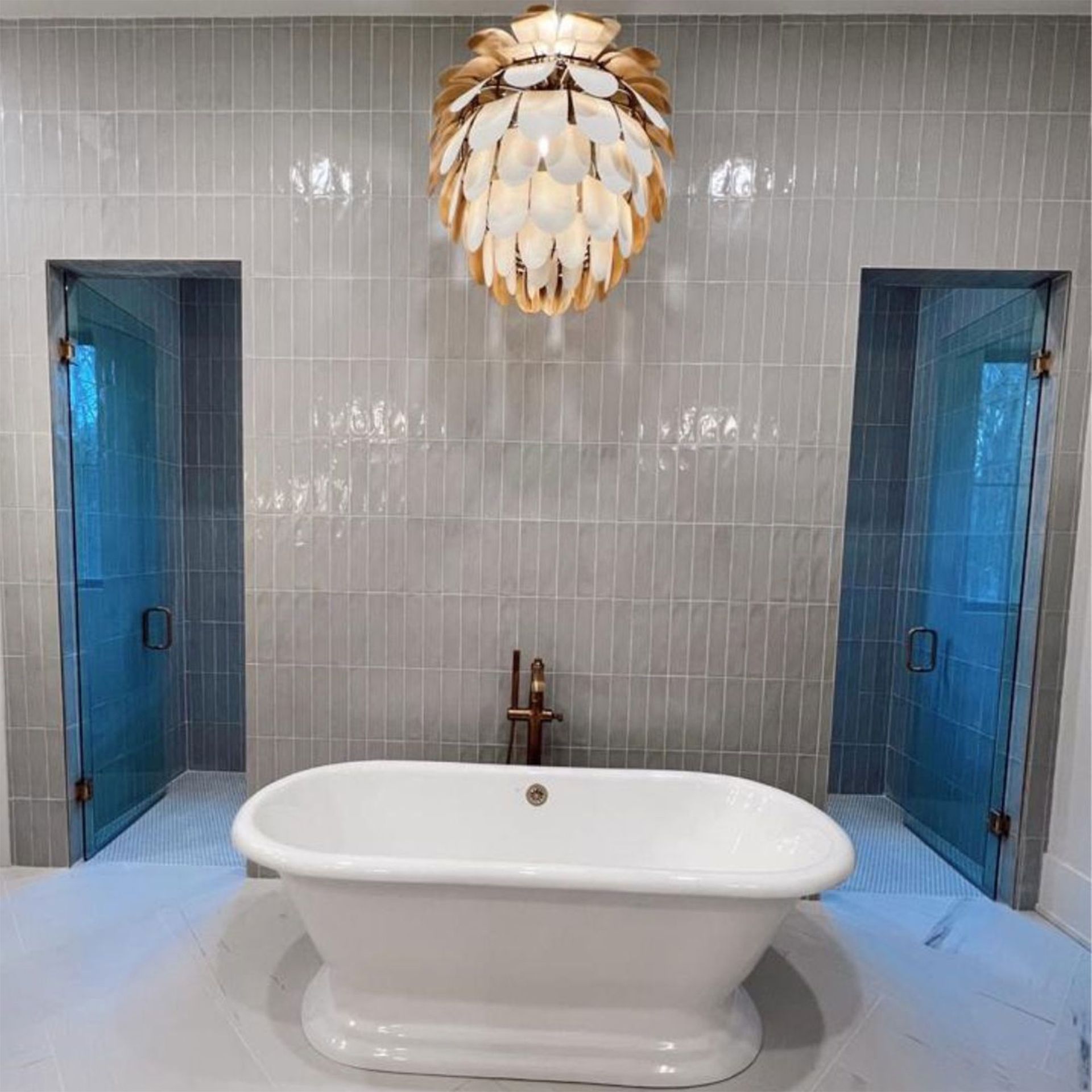 Bathroom with a white freestanding tub, two glass-door showers, and a gold pendant light. Gray tiled walls.