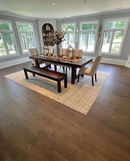 Dining room with large wood table, bench, and chairs, set on a patterned rug, surrounded by windows.