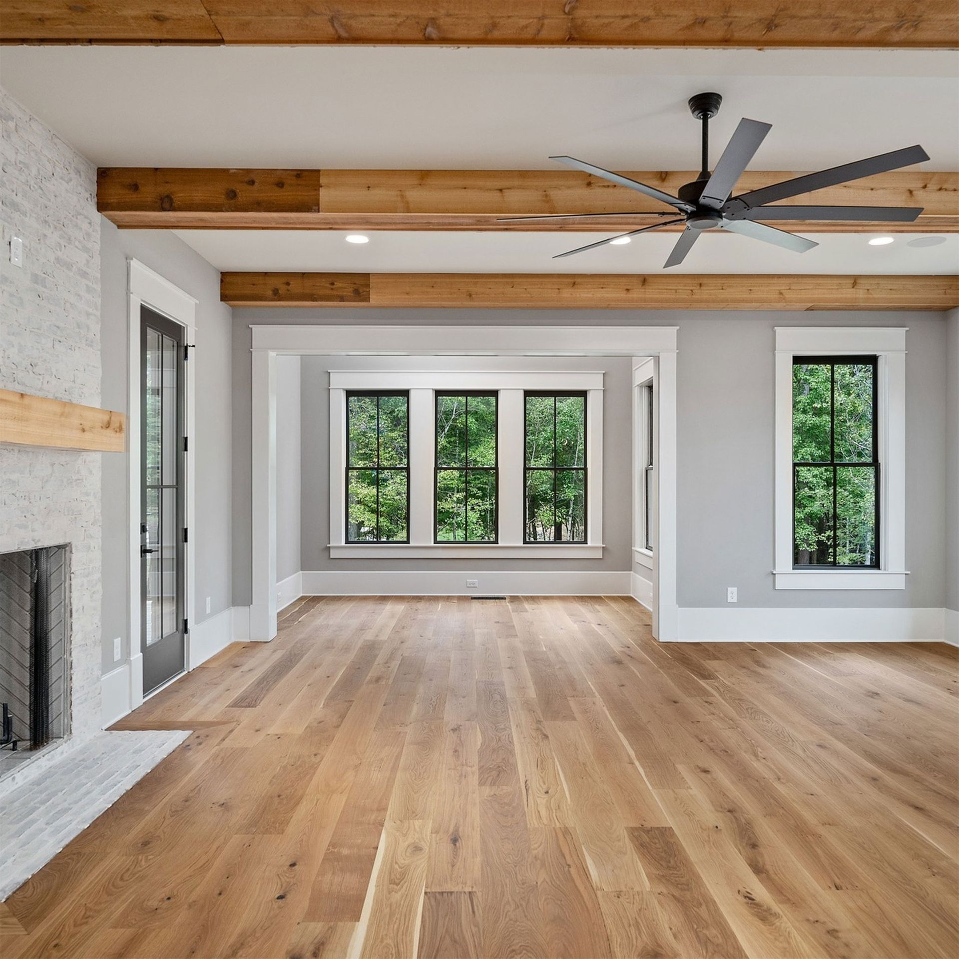 Spacious room with wood floors, exposed beams, and a fireplace. Large windows look out to green trees.