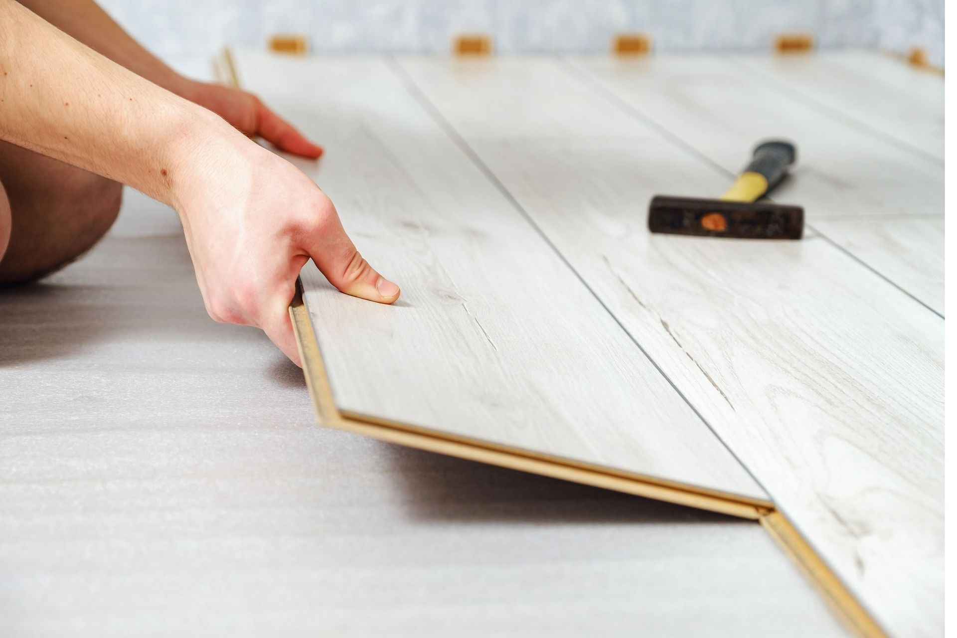 Person installing laminate flooring, light wood planks, hammer in background.
