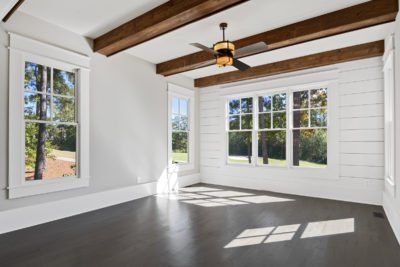 Empty room with dark floor, white walls and trim, wooden ceiling beams, and large windows with outdoor view.