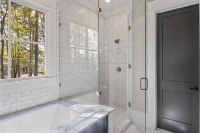 Bathroom with white tile, glass shower, black door, and a window overlooking trees.