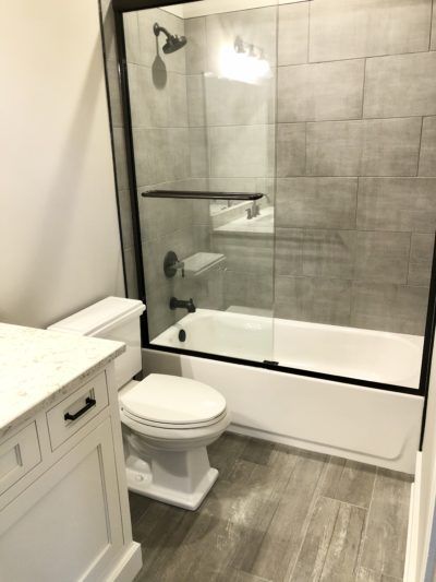 Bathroom with a white tub, toilet, and vanity. Gray tile and a glass shower door.