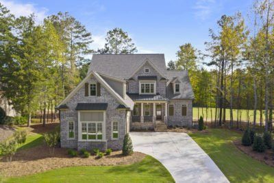 Two-story gray brick house with a driveway surrounded by trees and grass on a sunny day.