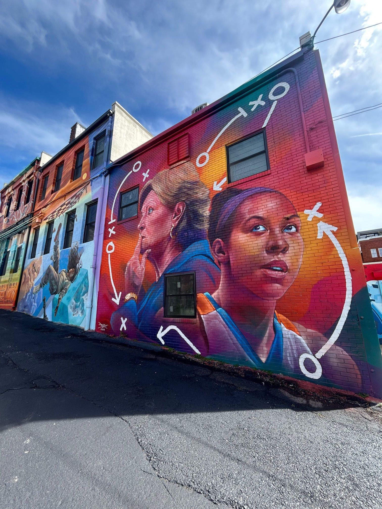 Mural on brick building, featuring portraits of two women with a basketball play design overlayed. Bright colors on a sunny day.