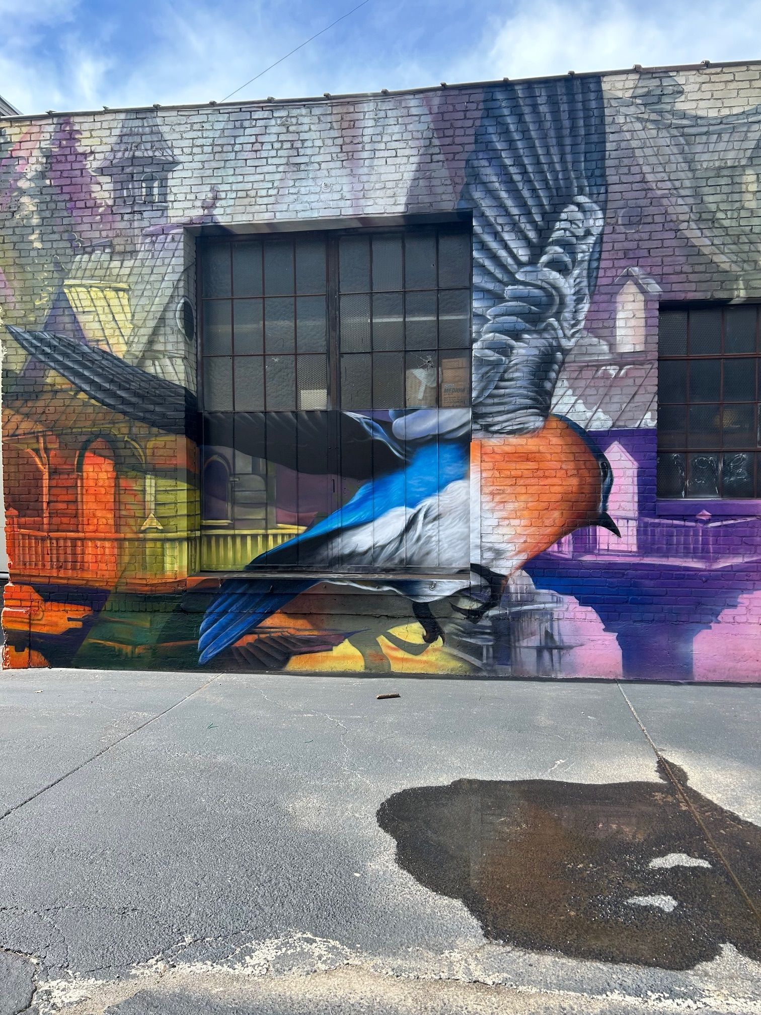 Mural of a bluebird in flight on a brick building. Background depicts a colorful city scene.