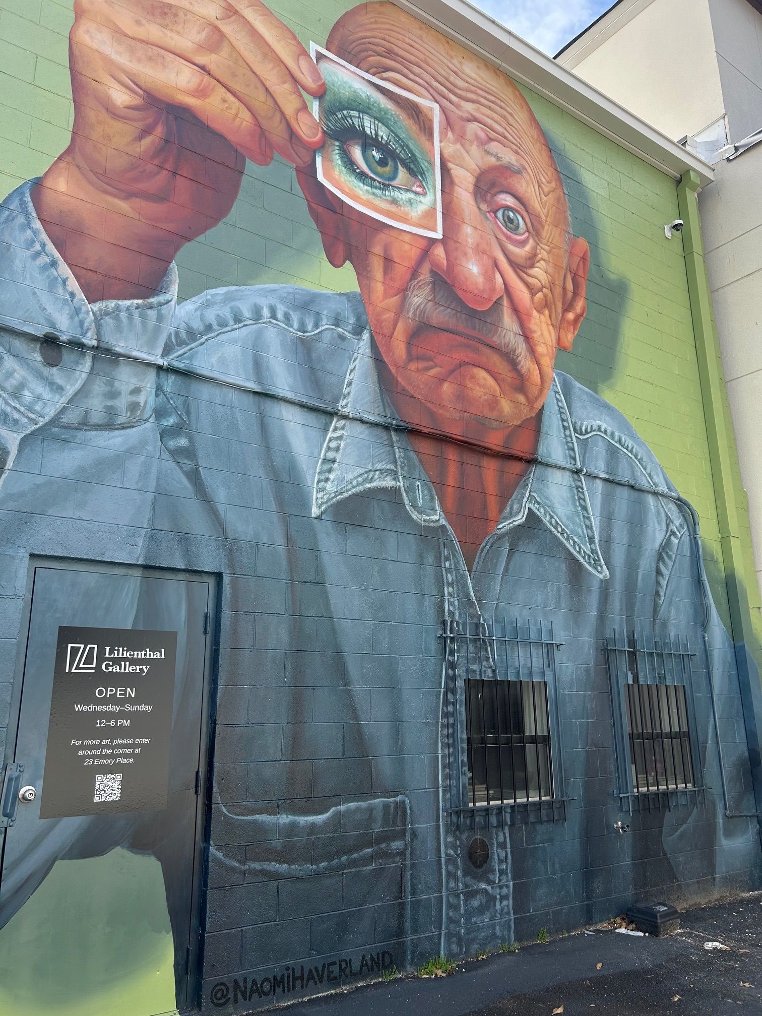 Mural of an elderly man holding a photo of an eye to his own, green and blue tones, outside a building.