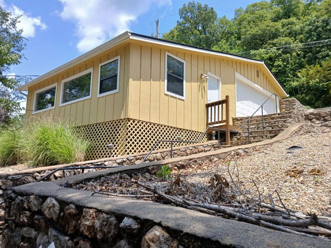 Small yellow house on a hillside with a wooden porch, lattice skirting, and rocky landscaping under a blue sky.