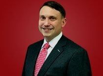 Man in a dark suit and pink tie smiling against a red background